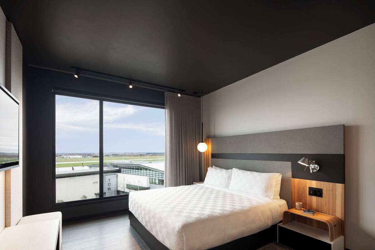A modern hotel room with a large window, a bed, a bedside table, and a nightstand.