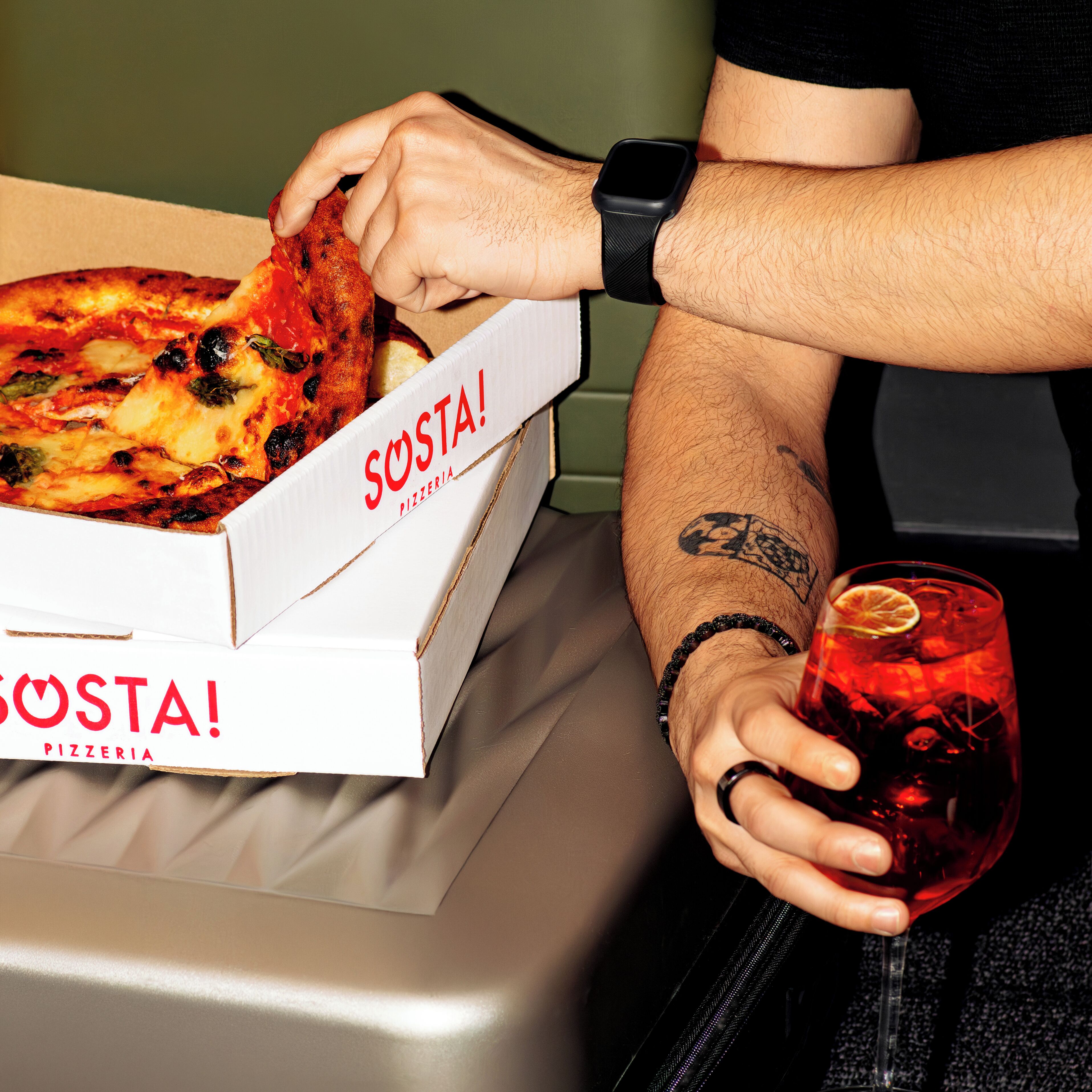 En person holder et pizzastykke og en drik, med en pizzabakke mærket "SOSTA PIZZERIA" på et bord.