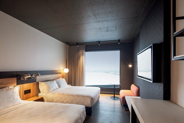 Habitación de hotel con dos camas, un escritorio, una silla, un ventanal con vistas y un televisor montado.