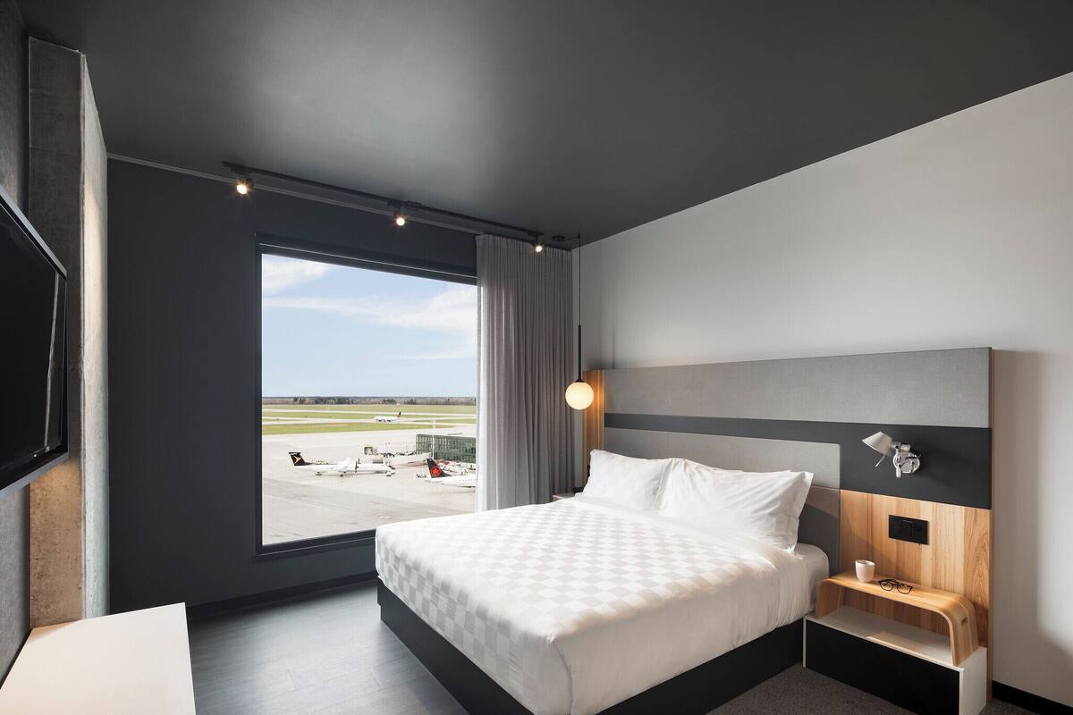 A modern hotel room with a large bed, a bedside table with a lamp, a TV, and a window with a view of an airport.