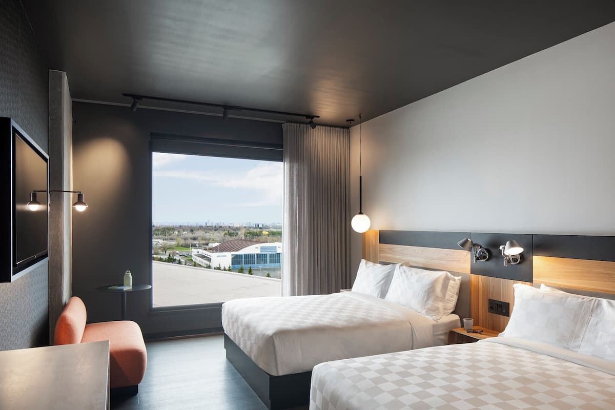 A modern hotel room with two beds, a TV, a small table, and a view of the city.