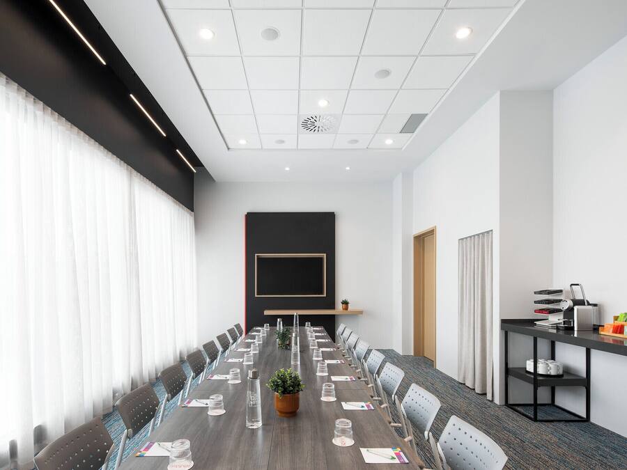 Un salón de conferencias con una larga mesa, sillas, una planta y una pantalla de proyector.