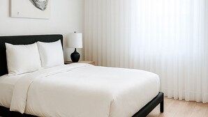Une chambre à coucher au style minimaliste, avec un lit, une table de chevet, une lampe et une grande fenêtre ornée de rideaux légers.