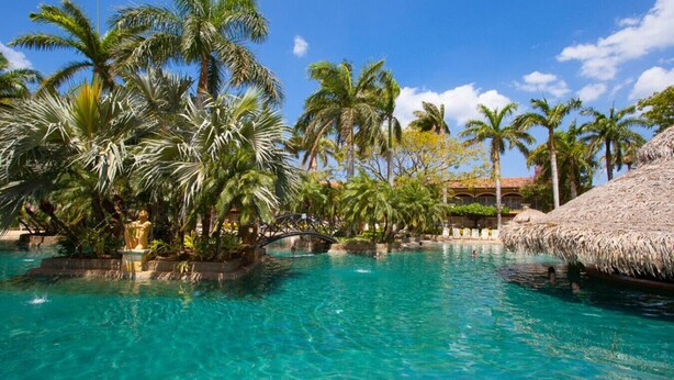 A tropical resort with clear blue water, palm trees, and a thatched-roof structure. - Tamarindo Diria 403 Luxury Ocean View Condo (Tamarindo)