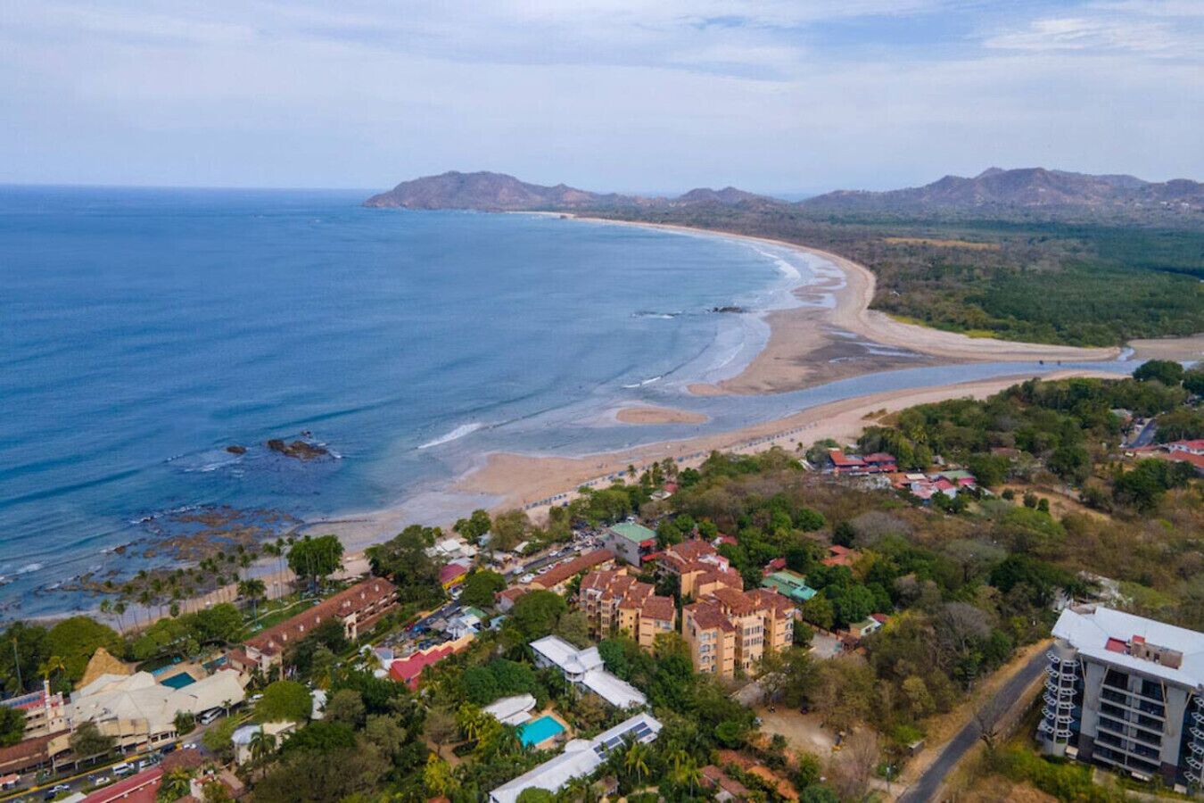 Vue aérienne d’une ville côtière avec une plage, des immeubles résidentiels et un paysage montagneux en arrière-plan.