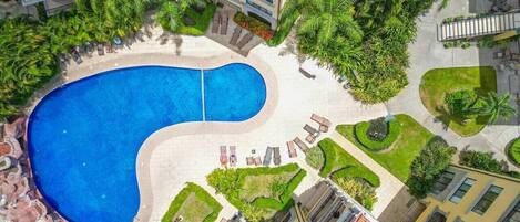 Vista aérea de un hotel con una amplia piscina, múltiples balcones y un exuberante paisaje.
