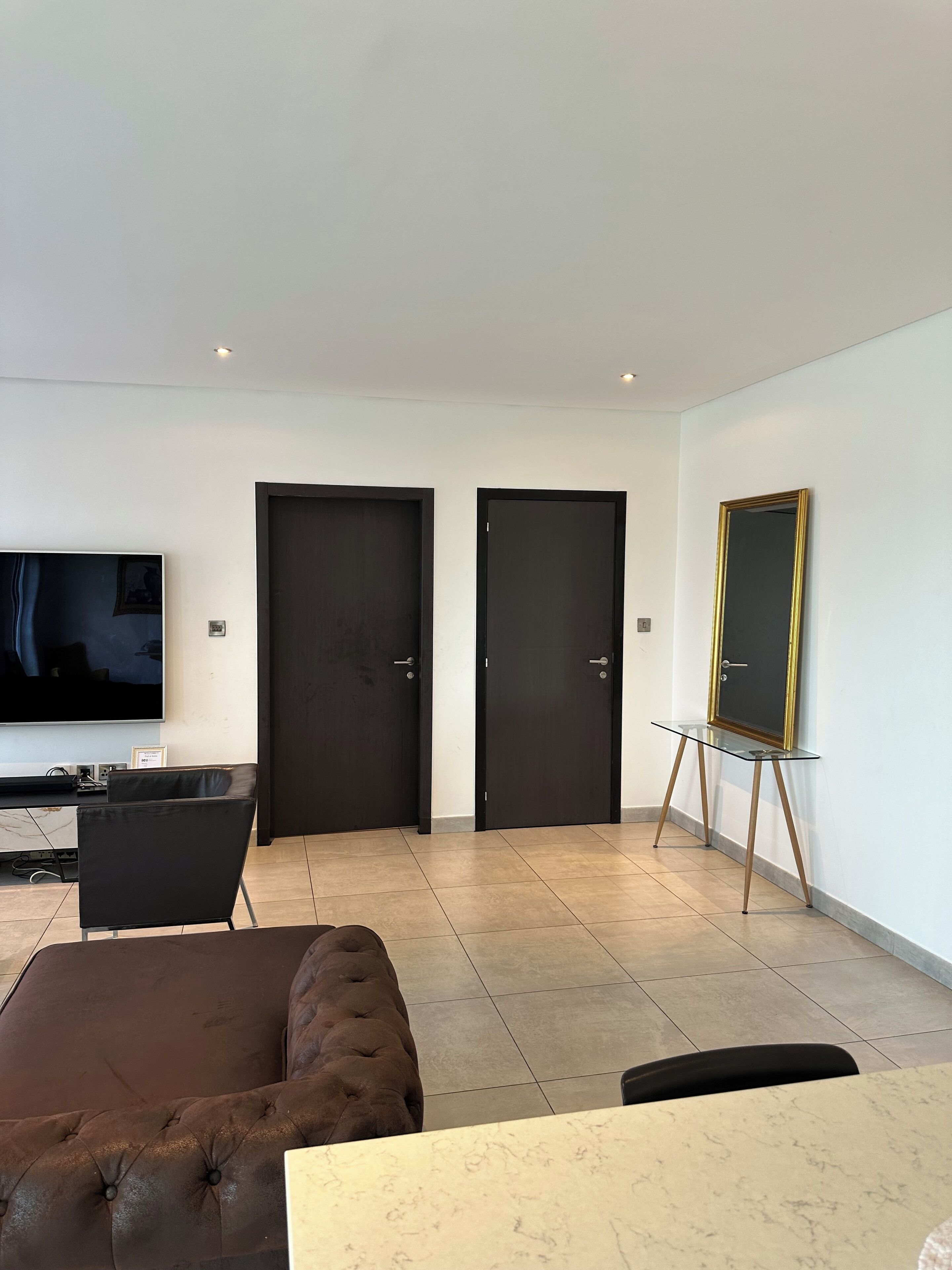 A modern living room with a large flat-screen TV, a brown tufted sofa, a glass coffee table, and two dark doors.