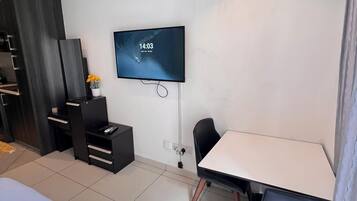 A hotel room with a bed, a desk, a chair, a TV, and an air conditioning unit.
