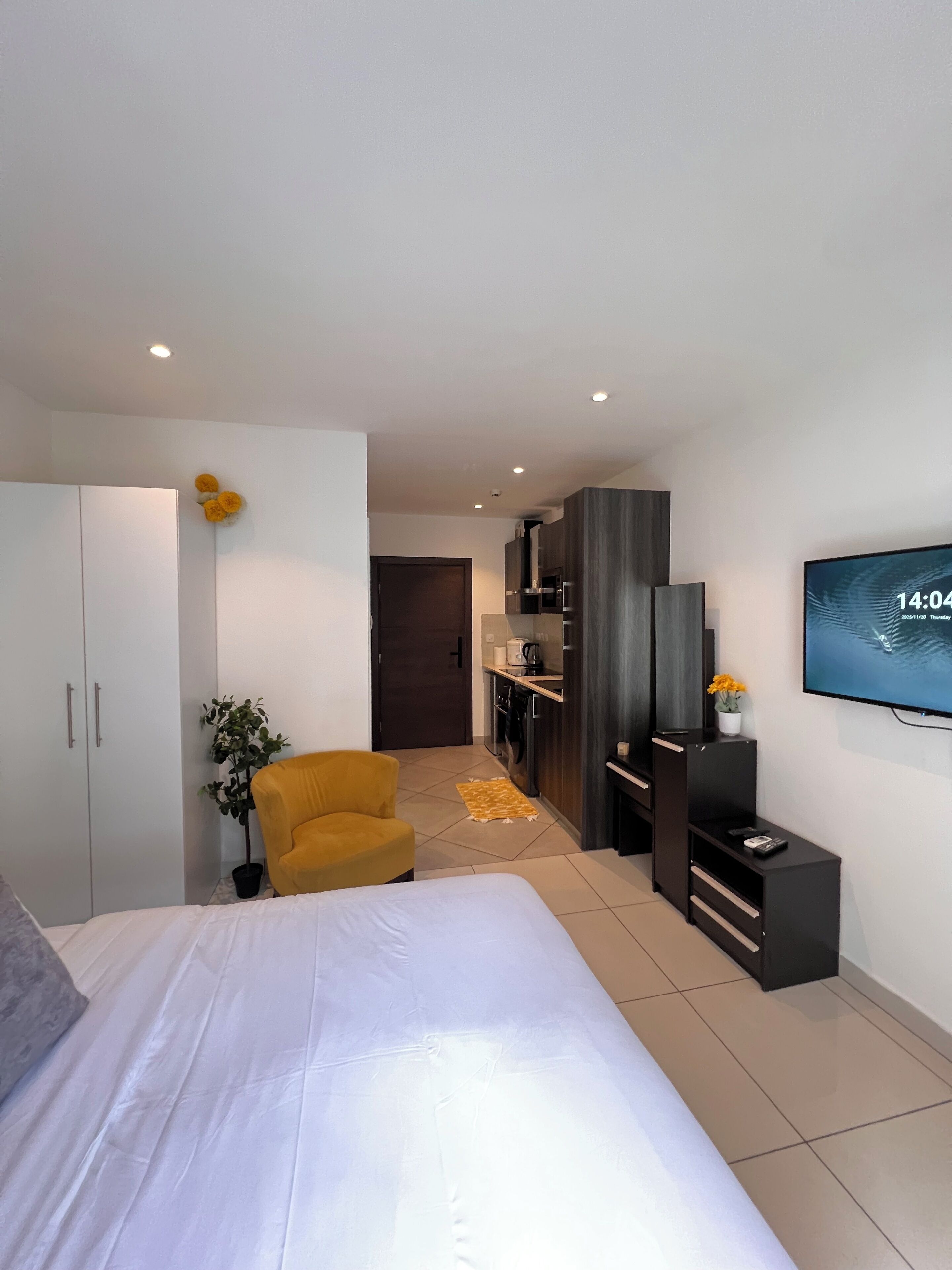 A modern bedroom with a bed, a yellow armchair, a TV, and a kitchenette in the background.