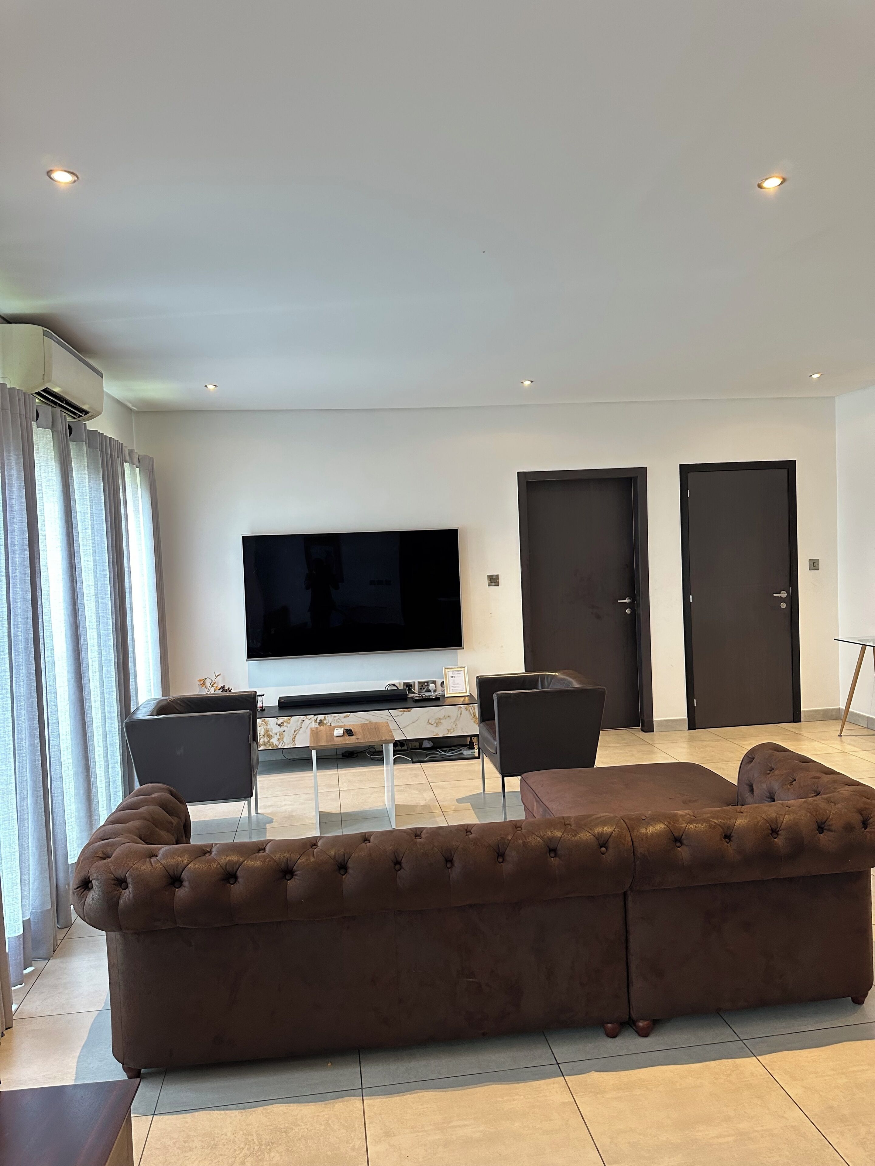 A modern living room with a large flat-screen TV, a brown sectional sofa, two armchairs, and a coffee table. There are two dark doors and a window with curtains.