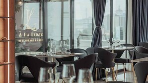 Interior de restaurante con mesas redondas, sillas negras y grandes ventanales con vista a la ciudad.