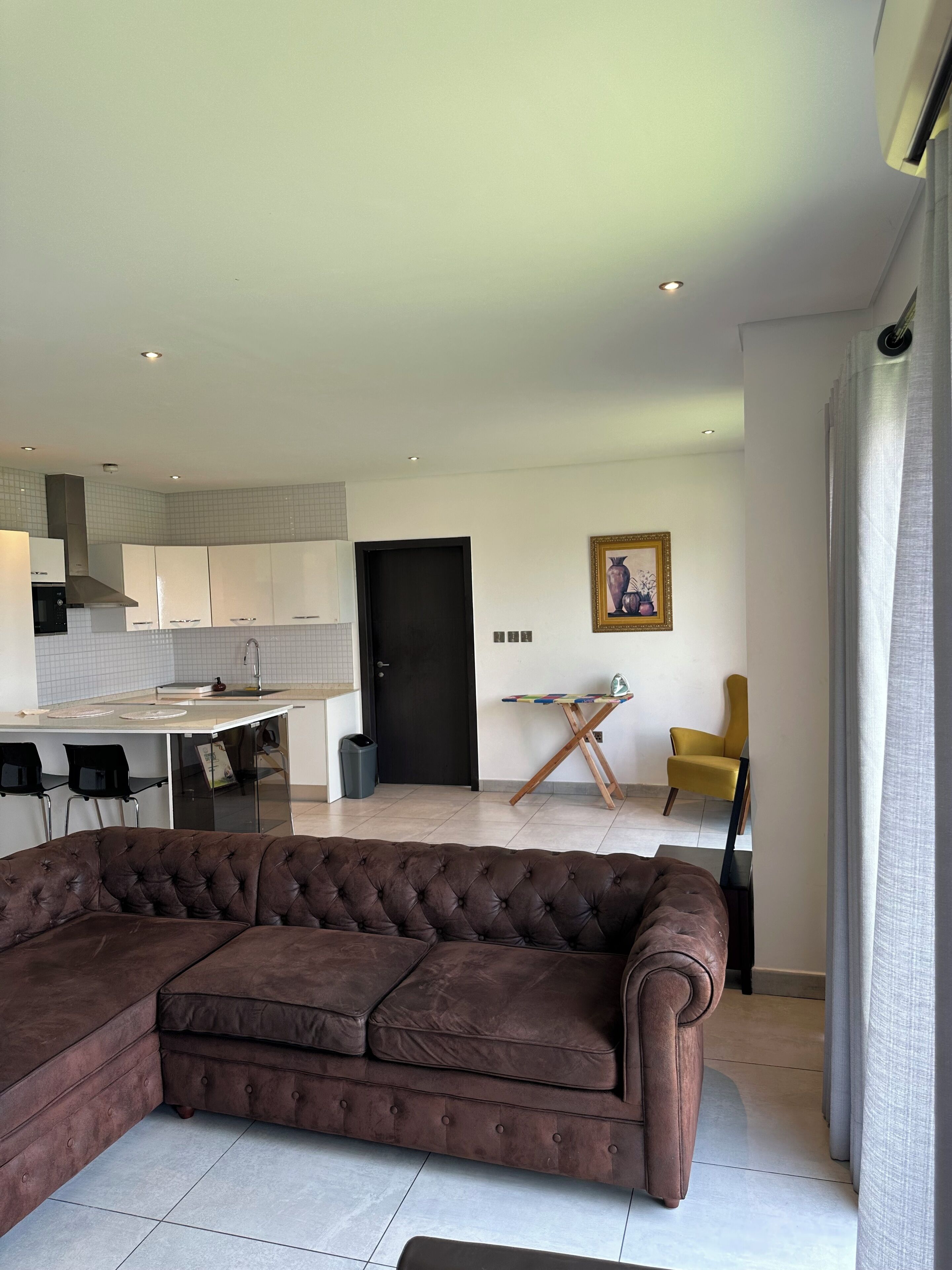 A modern living room with a brown sectional sofa, a dining area with a small table and chairs, and a kitchen with white cabinets and a black door.