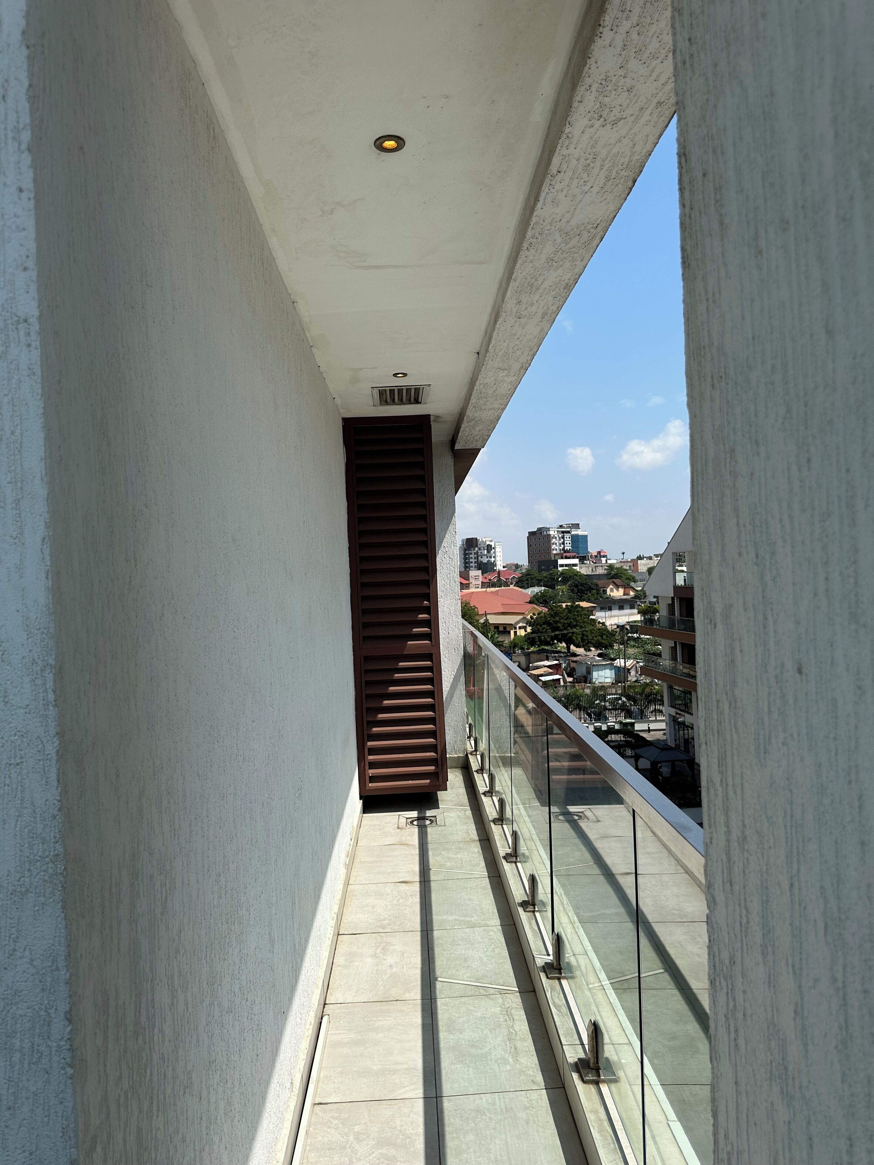 Un balcon étroit doté d’une rambarde en métal, une fenêtre à persiennes marron et une vue dégagée sur un paysage urbain avec des bâtiments et un ciel bleu.