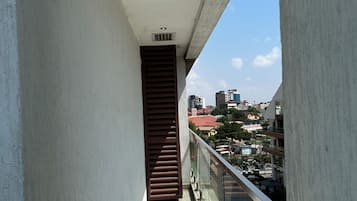 Un balcon étroit doté d’une rambarde en métal, une fenêtre à persiennes marron et une vue dégagée sur un paysage urbain avec des bâtiments et un ciel bleu.