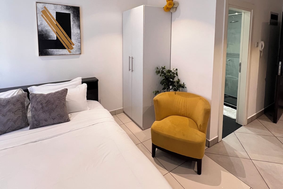 Un dormitorio con cama, armario, sillón amarillo, planta en maceta y baño a la vista a través de una puerta abierta.