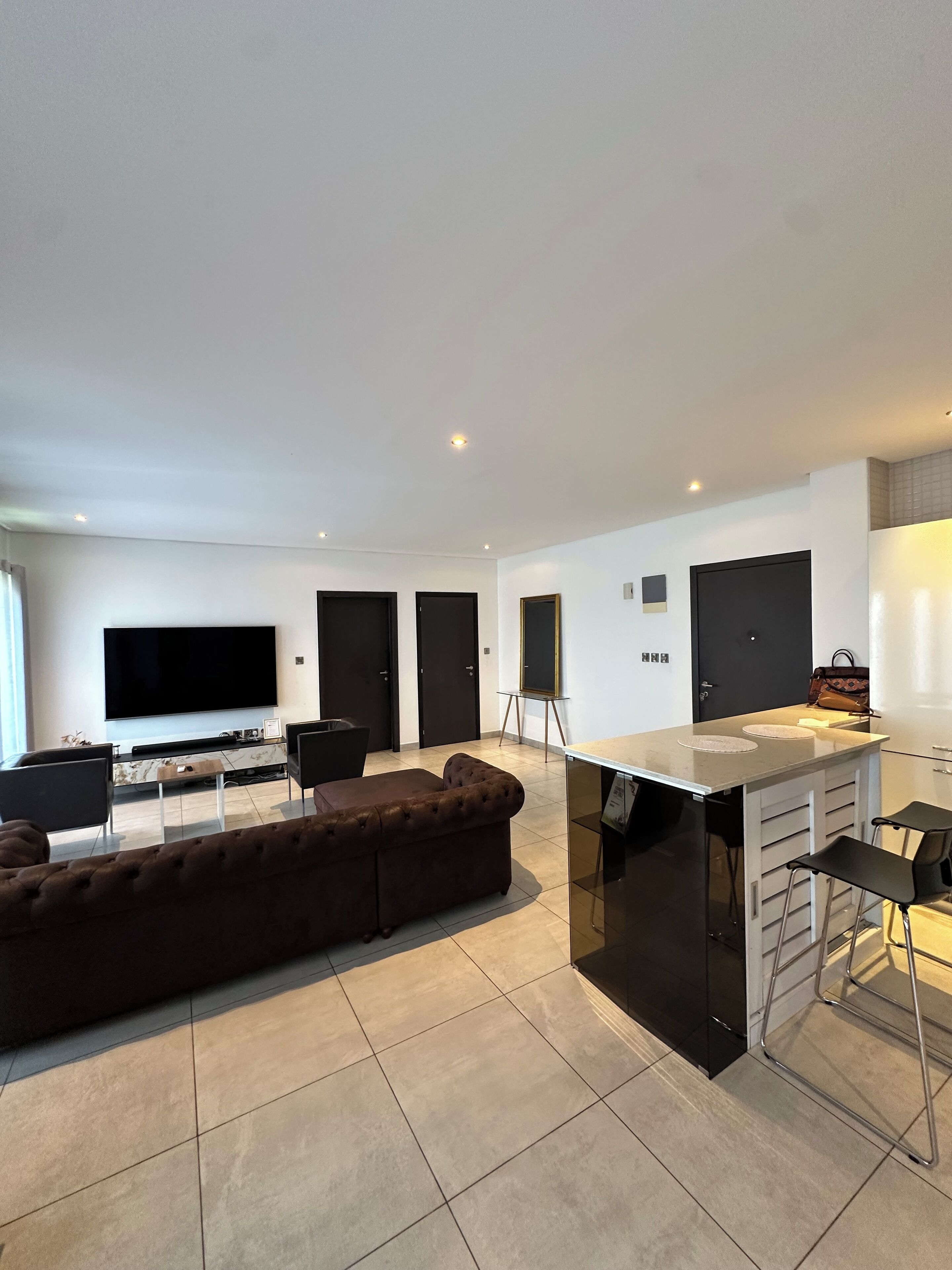A modern living room with a large flat-screen TV, a brown sectional sofa, a small coffee table, and a kitchen area with a white countertop and bar stools.