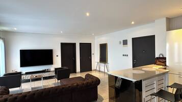 A modern living room with a large flat-screen TV, a brown sectional sofa, a small coffee table, and a kitchen area with a white countertop and bar stools.