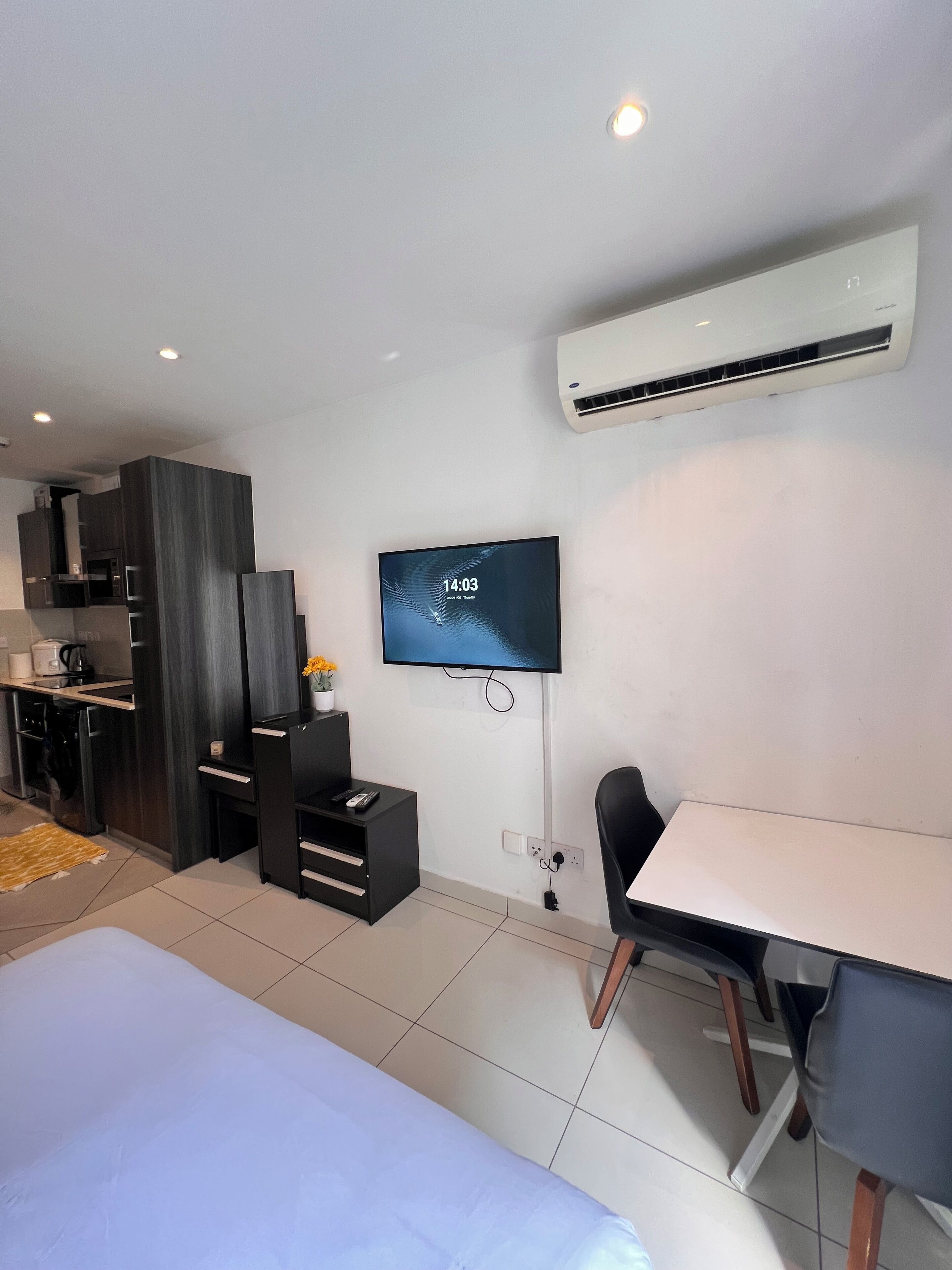 A modern hotel room with a bed, a TV mounted on the wall, a desk with a chair, a kitchenette, and an air conditioning unit.