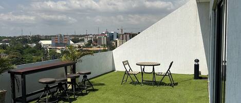 A rooftop terrace with artificial grass, a wooden table and chairs, and a view of the city skyline.