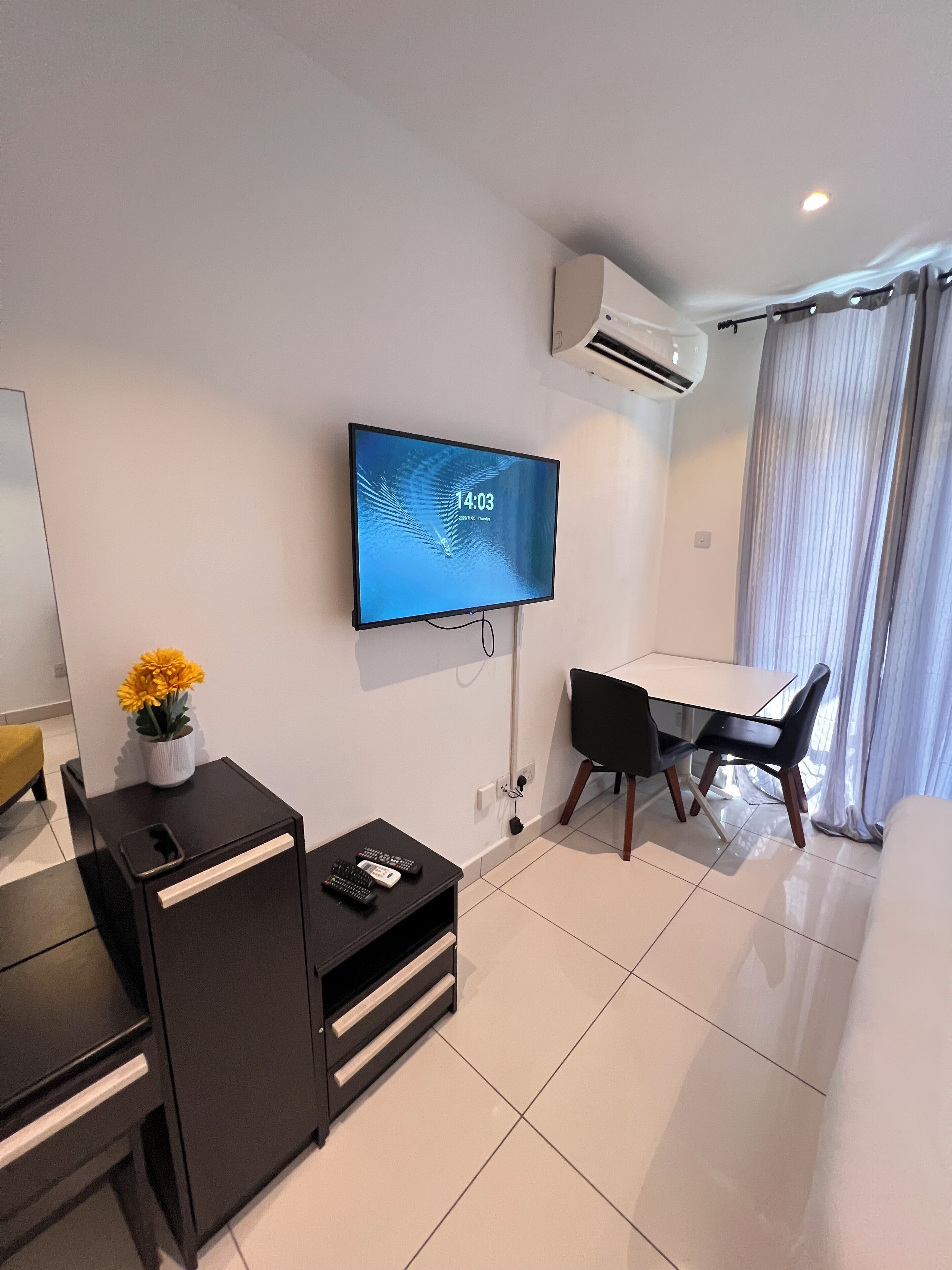 A modern hotel room with a flat-screen TV mounted on the wall, a small desk with a chair, a bedside table with a vase of flowers, and a large window with curtains.