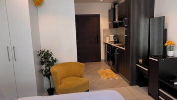 Un dormitorio moderno con una cama grande, un sillón amarillo, una pequeña planta en maceta y una kitchenette al fondo.