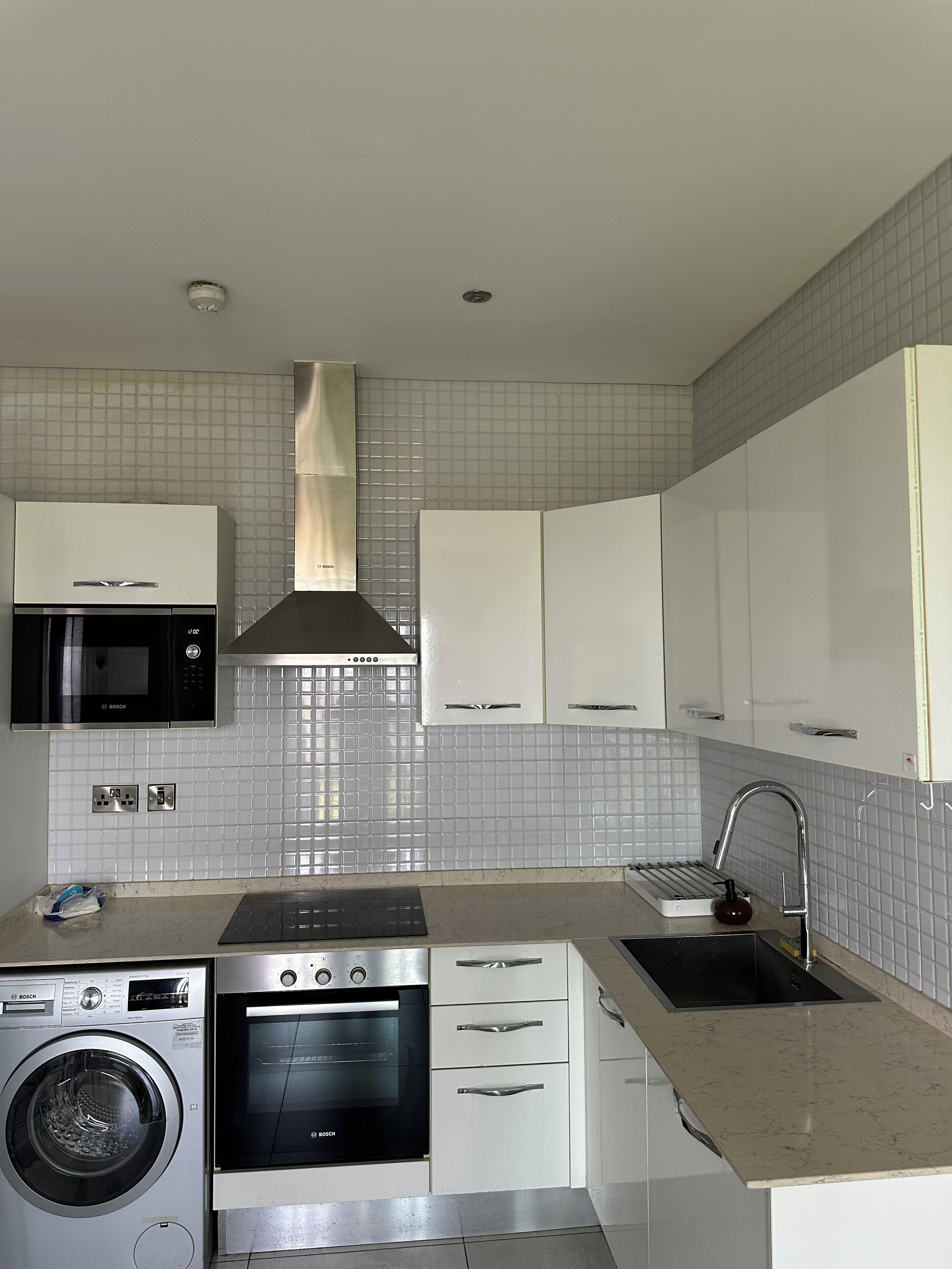 A modern kitchen with white cabinets, a Bosch microwave, oven, and washing machine, a stainless steel sink, and a tiled backsplash.