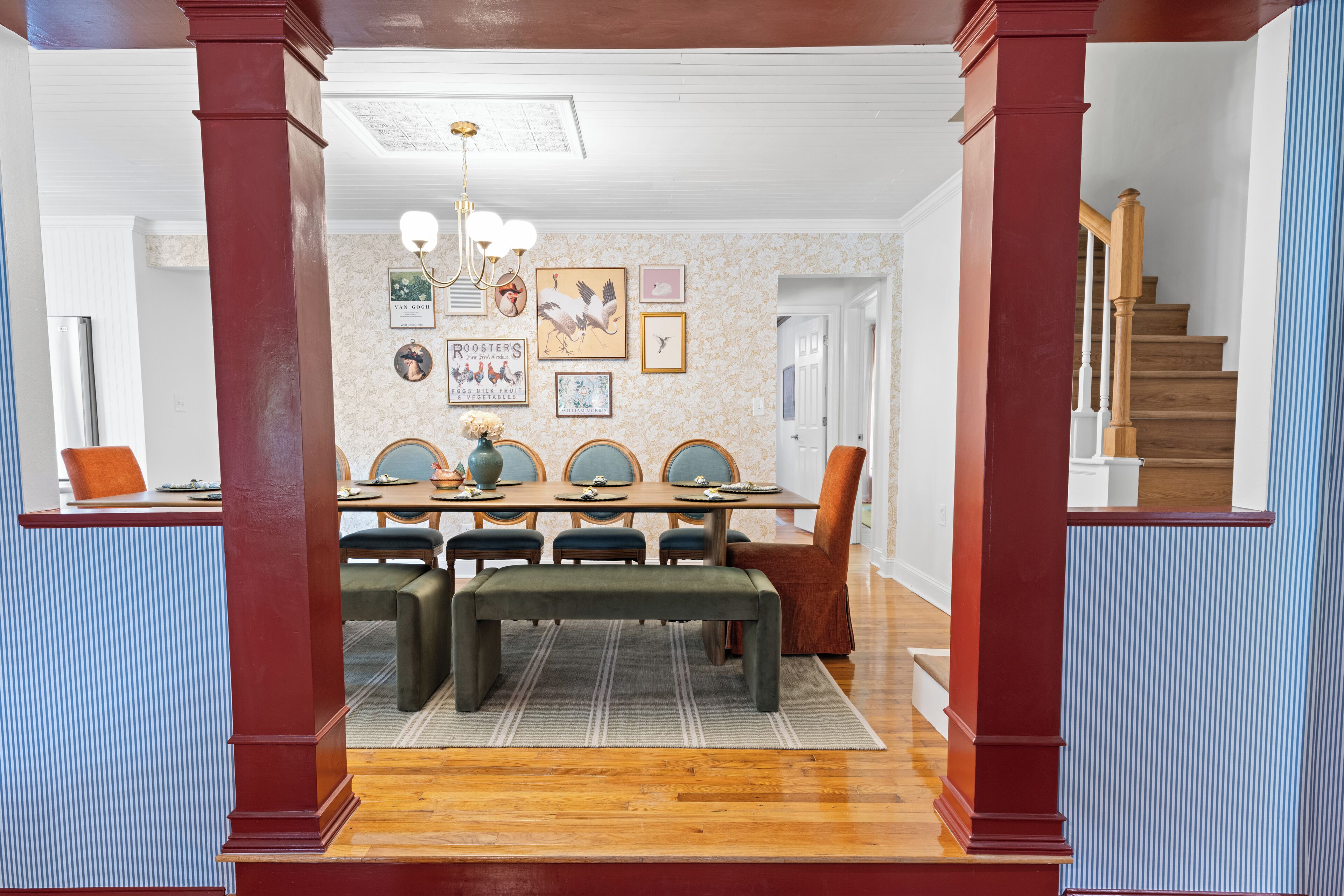 Un comedor con una mesa de madera, sillas y un banco. Hay cuadros en la pared y una lámpara de araña sobre la mesa.