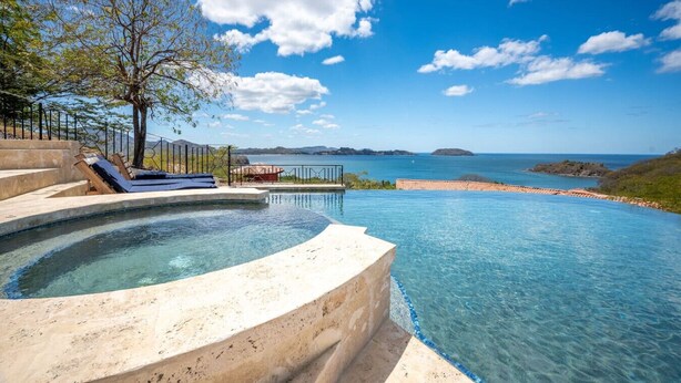 A infinity pool with clear blue water, a tree, and a view of the sea and distant islands. - Luxury Living at Villas Catalina #13 (Potrero)