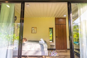 A bedroom with a bed, a small stool, a window with a view of greenery, and a door. - Moani Punta Uva Lodge 5 min Walk to beach (Talamanca)