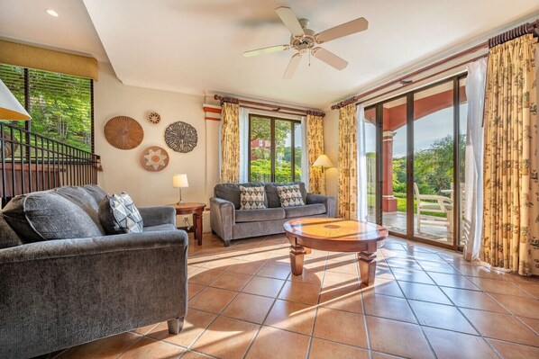 A spacious living room with a grey sofa, a round wooden coffee table, and a grey armchair. There is a ceiling fan, a window with curtains, and a view of greenery outside. - Sunshine Getaway- Lovely Golf Front Villa (Brasilito)