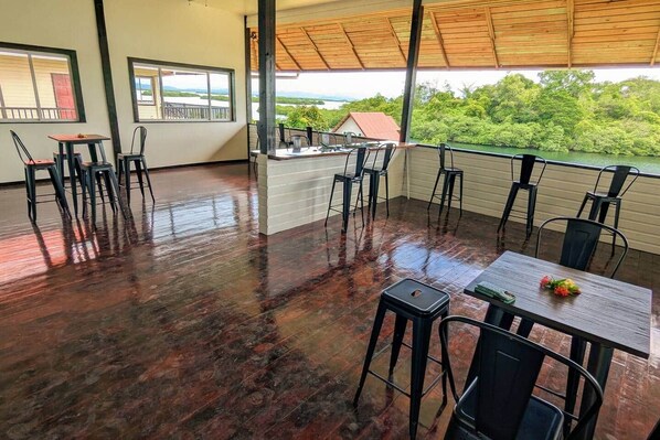 A covered outdoor seating area with tables and chairs, a wooden floor, and a view of greenery and a house in the background.