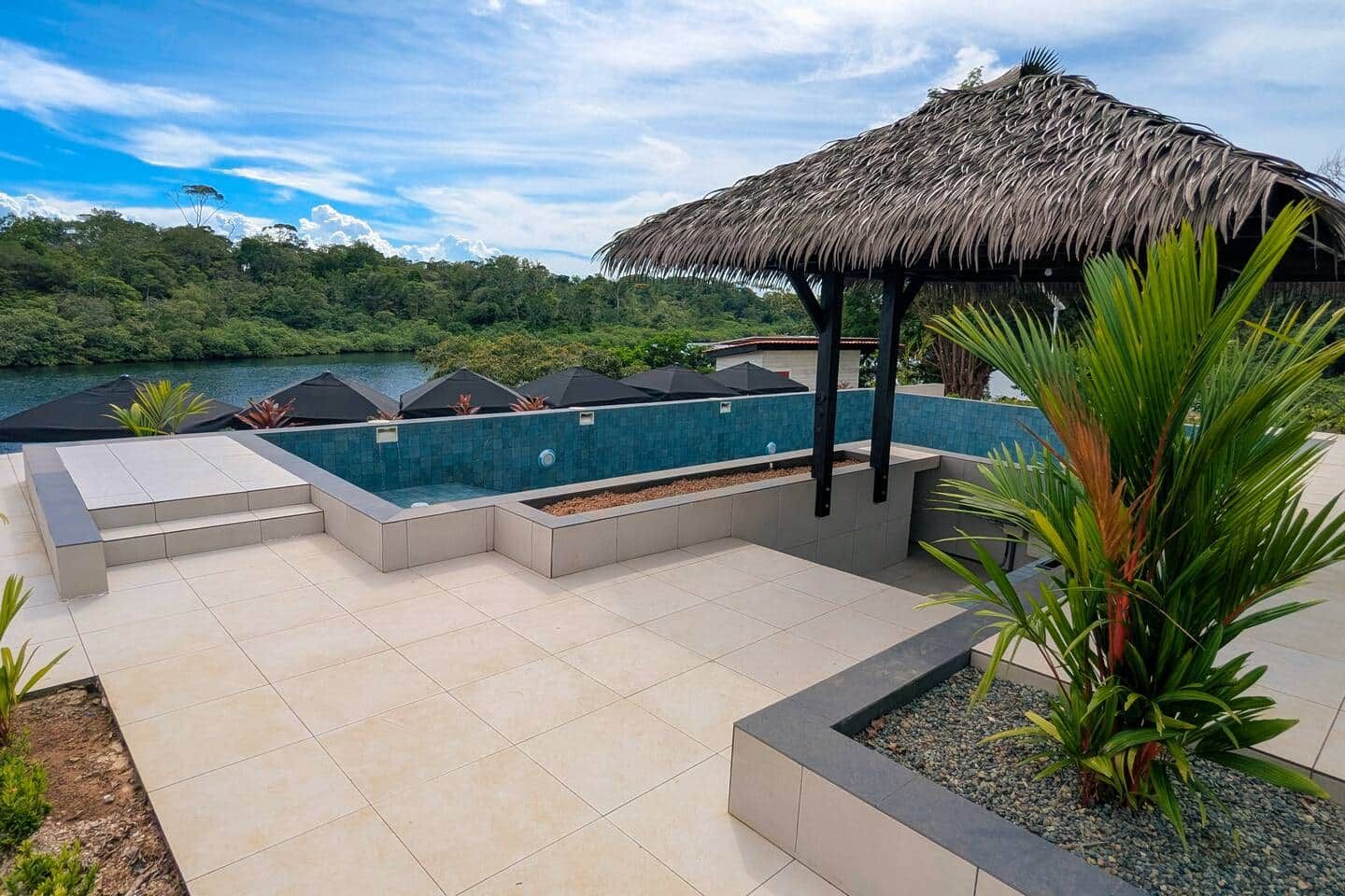 A swimming pool with a thatched roof structure, surrounded by a tiled deck and a view of a lake and forest.