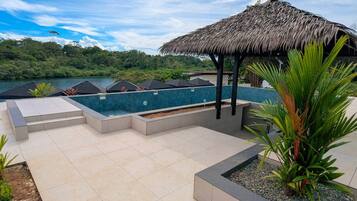 A swimming pool with a thatched roof structure, surrounded by a tiled deck and a view of a lake and forest.
