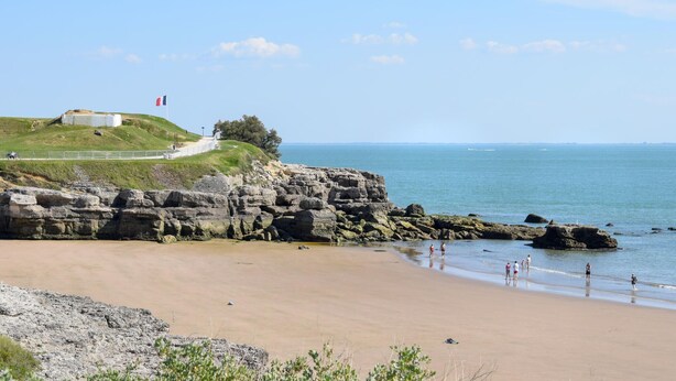 A coastal landscape with a sandy beach, rocky cliffs, and a grassy hill with a structure on top. - Appartement Proche Plage (Royan)