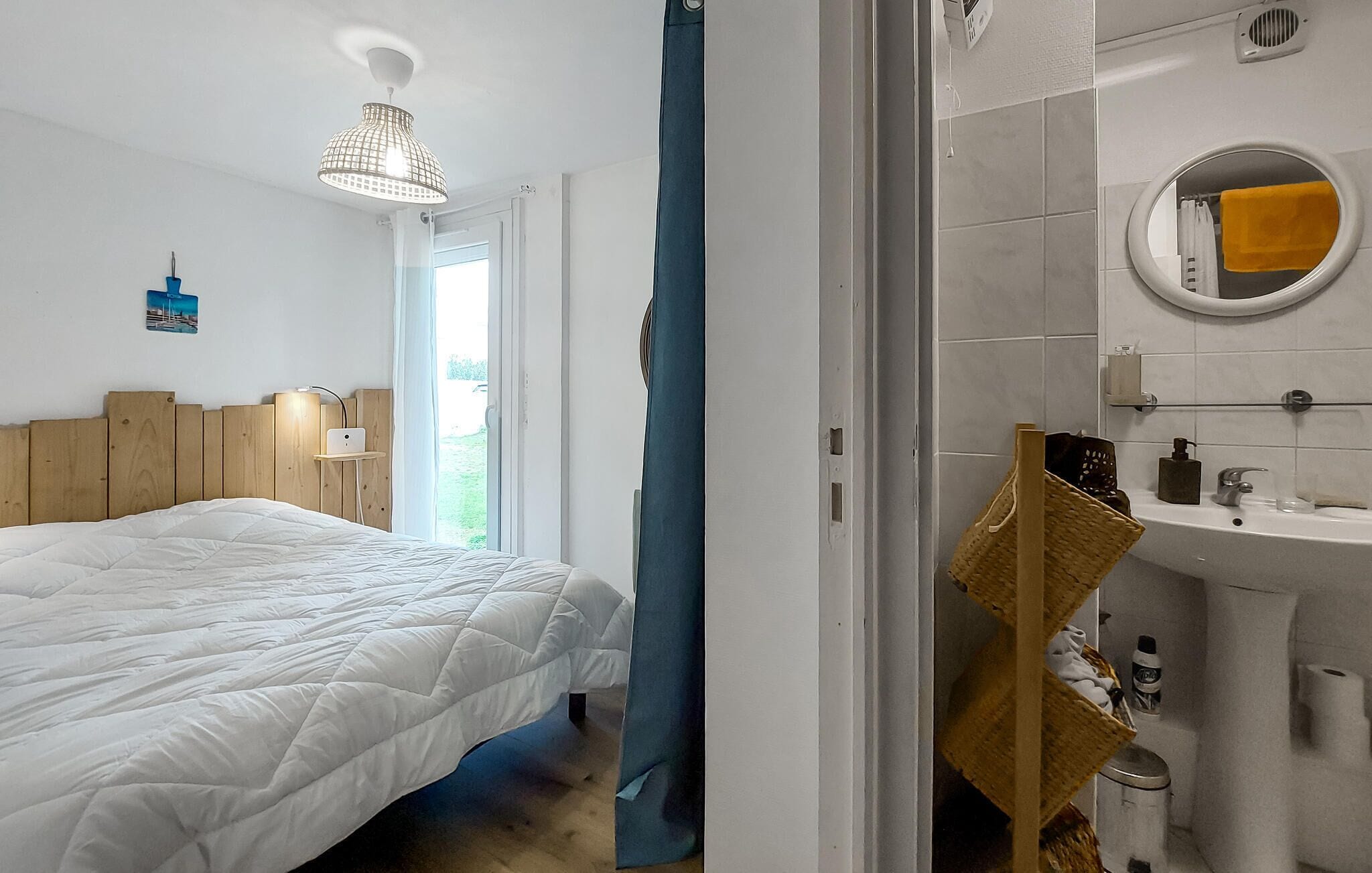 A bedroom with a bed, a bedside table with a lamp, a window, and a bathroom with a sink and mirror.