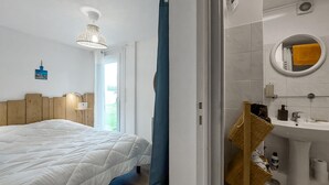 A bedroom with a bed, a bedside table with a lamp, a window, and a bathroom with a sink and mirror. - Appartement Proche Plage (Royan)