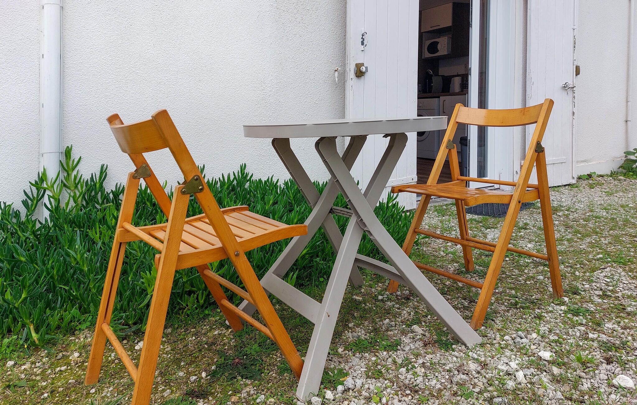 Deux chaises en bois et une table sont placées à l’extérieur, devant un bâtiment à la façade blanche.