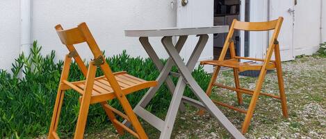 Deux chaises en bois et une table sont placées à l’extérieur, devant un bâtiment à la façade blanche.