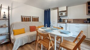 A compact living space with a sofa, dining table, and chairs, a kitchenette, and a small bookshelf. - Appartement Proche Plage (Royan)