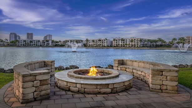 A circular stone fire pit with seating on a paved area by a body of water, with buildings and palm trees in the background.