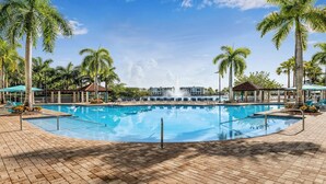 A large outdoor swimming pool with a fountain, surrounded by palm trees and lounge chairs.