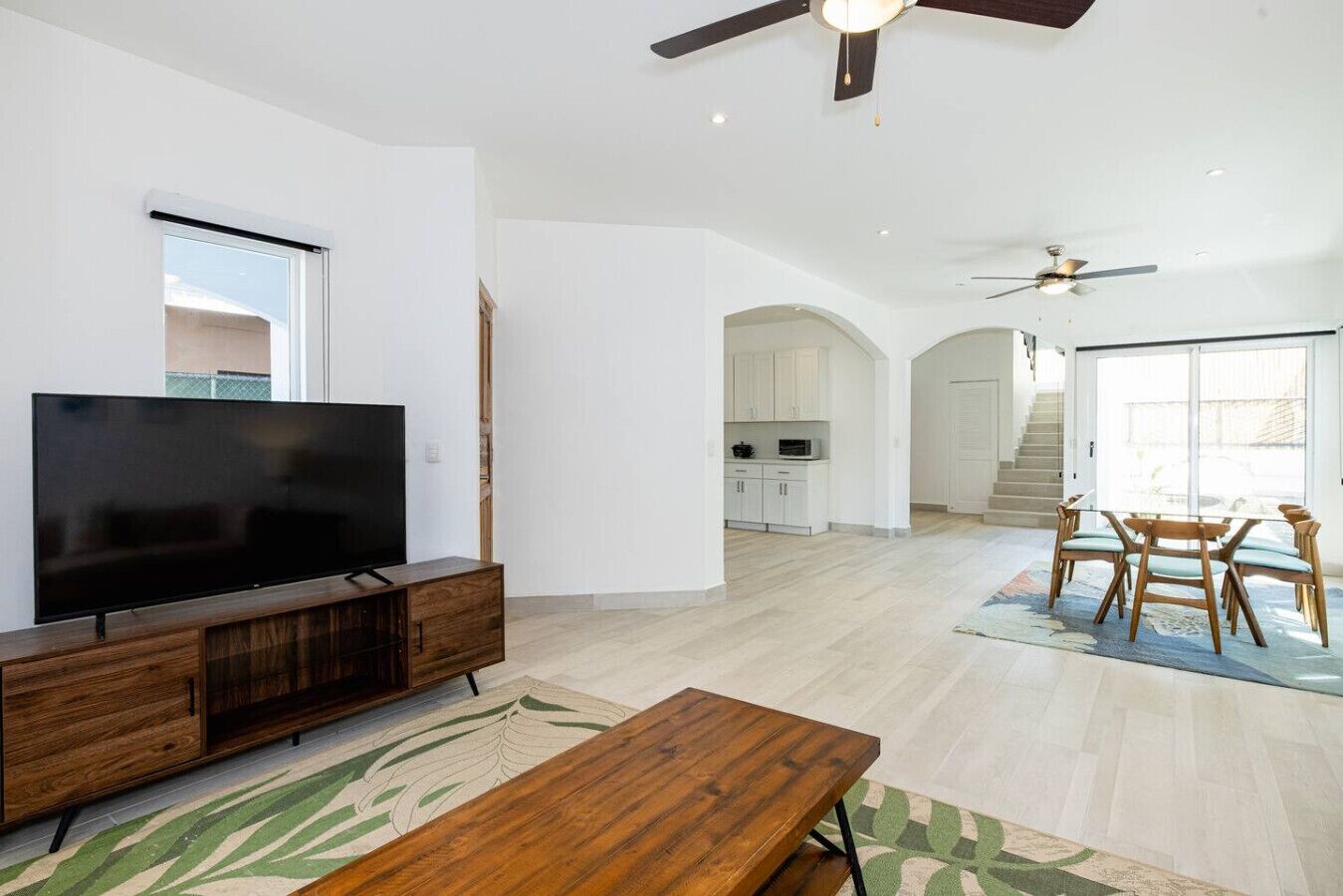 Un vaste salon doté d’un téléviseur à écran plat posé sur un meuble en bois, un coin repas avec une table et des chaises, et une cuisine à l’arrière-plan.