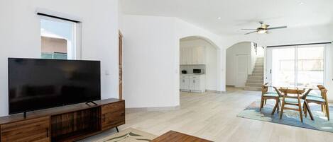 Un vaste salon doté d’un téléviseur à écran plat posé sur un meuble en bois, un coin repas avec une table et des chaises, et une cuisine à l’arrière-plan.