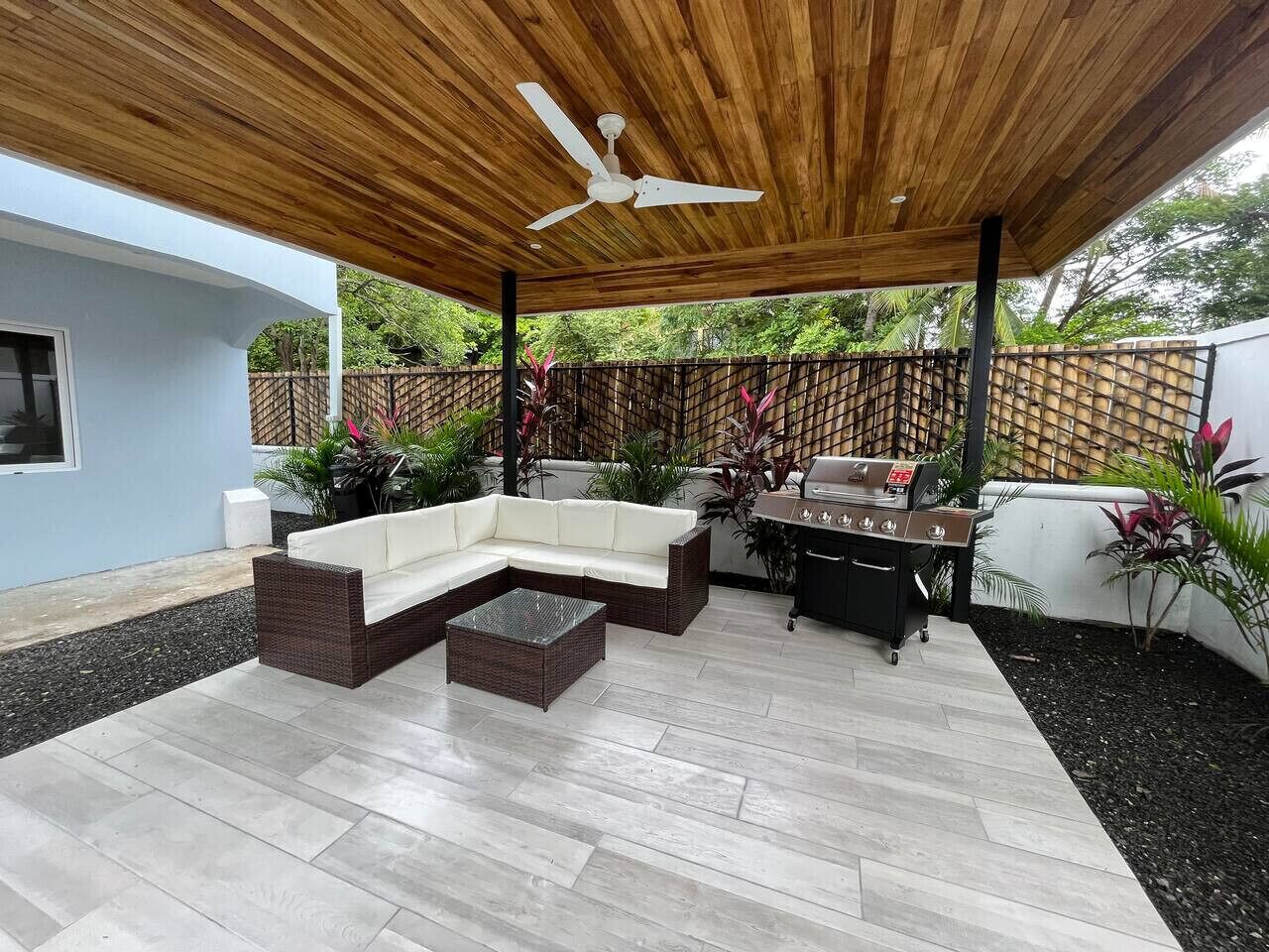 An outdoor patio with a wooden ceiling, a white ceiling fan, a white sofa, a wicker coffee table, and a barbecue grill.