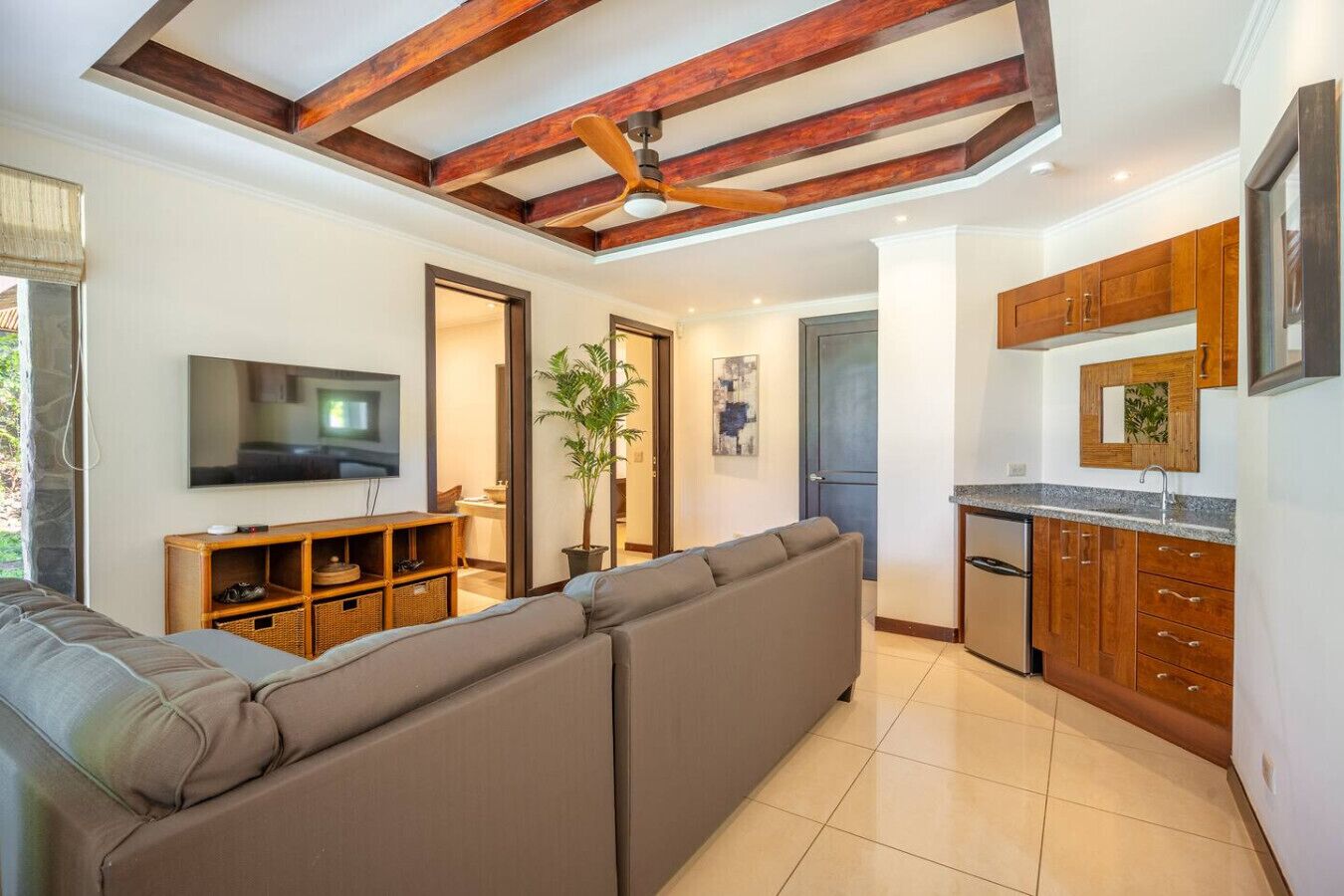 A modern living room with a sectional sofa, a flat-screen TV, wooden cabinets, and a tiled floor.