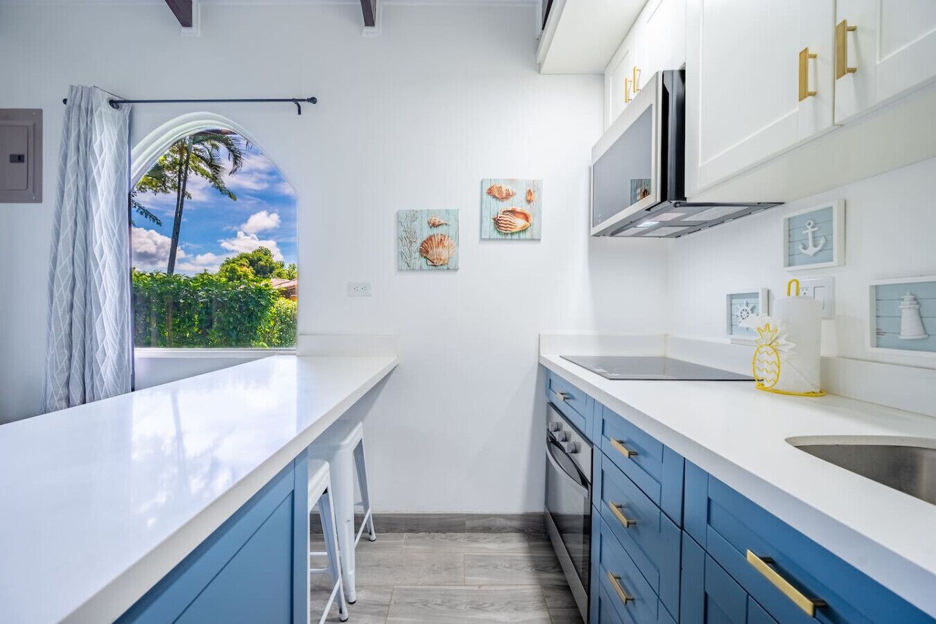 Cocina moderna con encimeras blancas, armarios azules y ventana con vistas a zonas verdes.