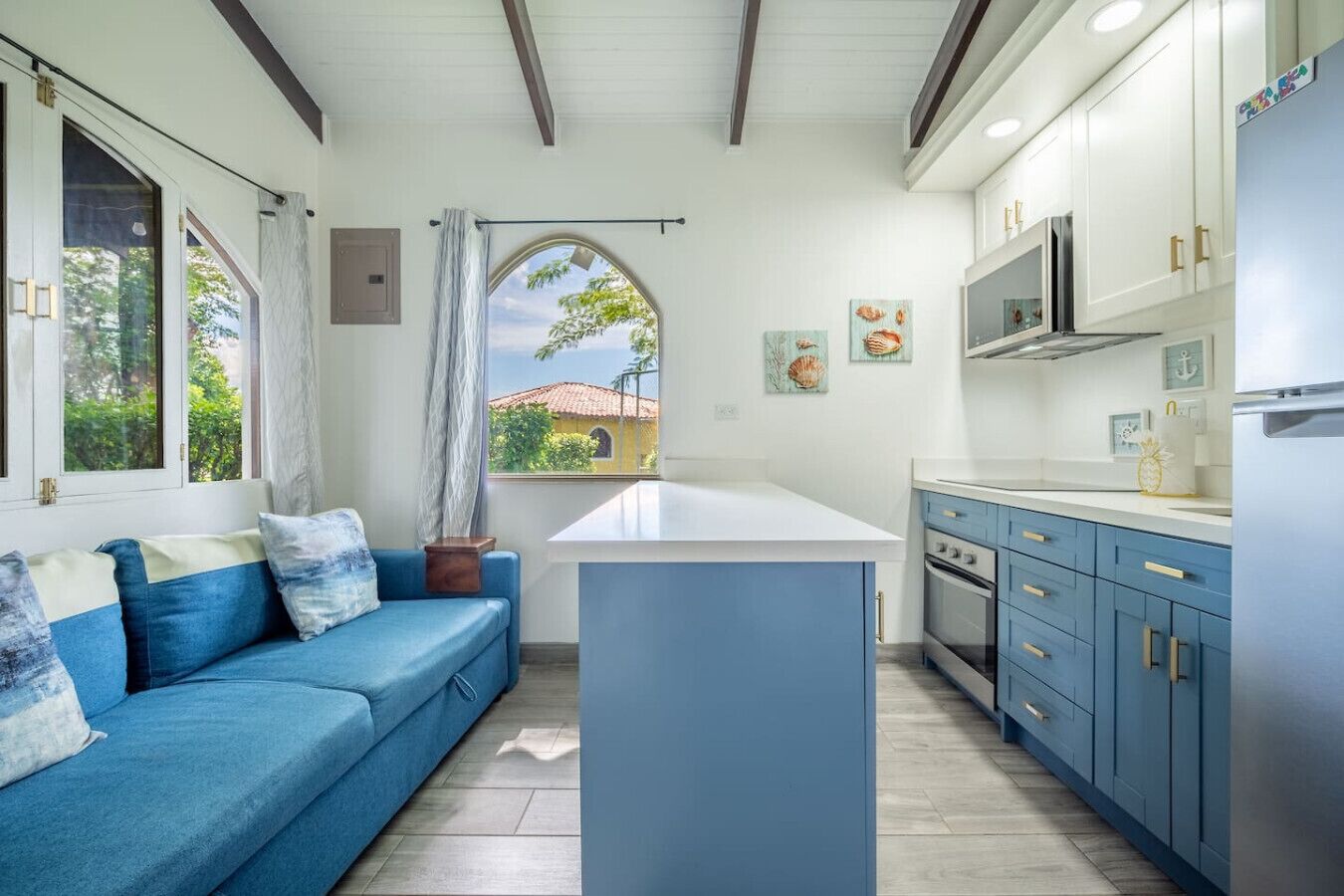 A modern kitchen with a blue sofa, a large island, and a view of a house through a window.