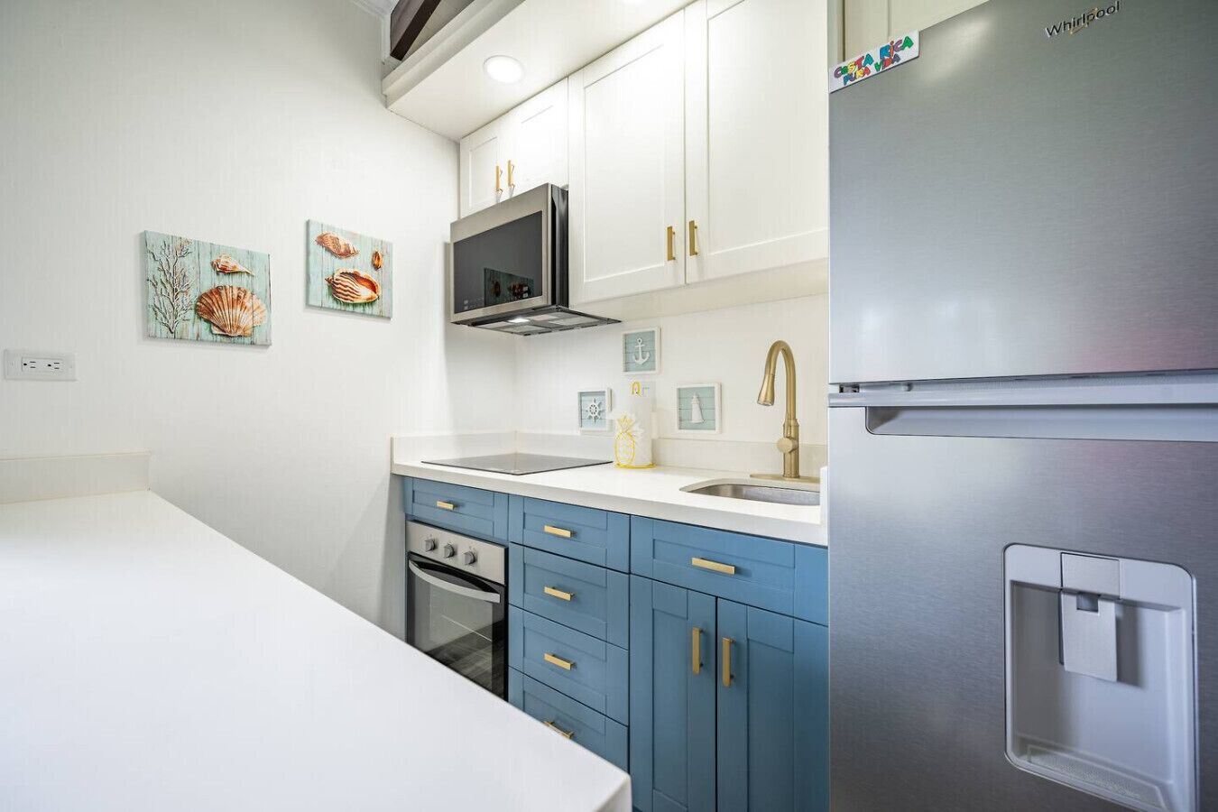 A modern kitchen with white cabinets, a stainless steel refrigerator, a microwave, and a sink with a gold faucet. There are decorative wall art pieces and a small oven.