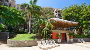 A resort with a thatched-roof structure, palm trees, and lounge chairs. - La Diria #303 Best Location, Walk to Beach/Shops! (Tamarindo)