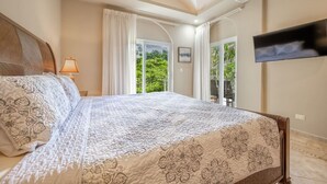 A bedroom with a large bed, a TV mounted on the wall, and a balcony with a view of greenery. - La Diria #303 Best Location, Walk to Beach/Shops! (Tamarindo)